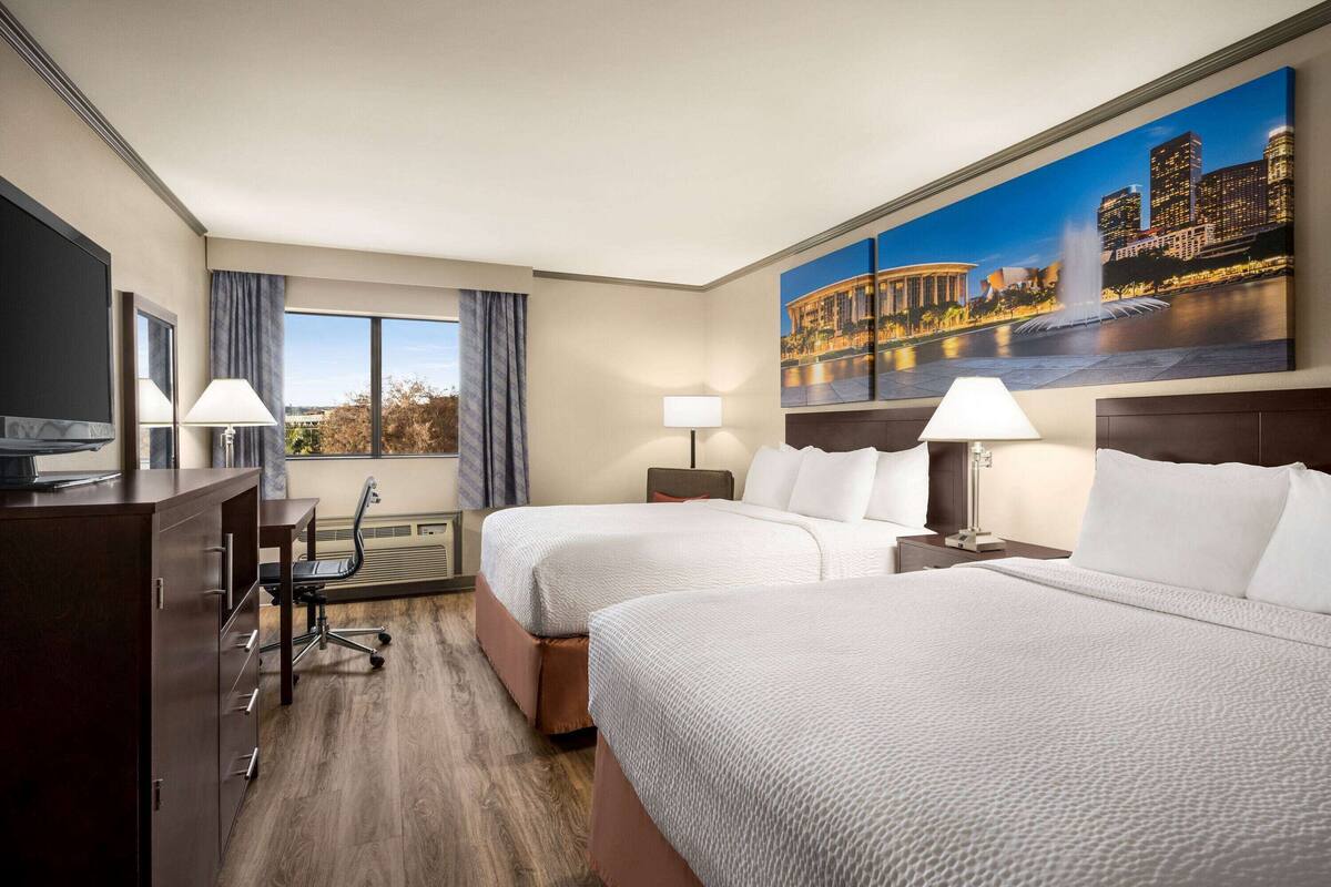 A hotel room with two beds, a desk, a chair, a TV, and a large window with curtains.