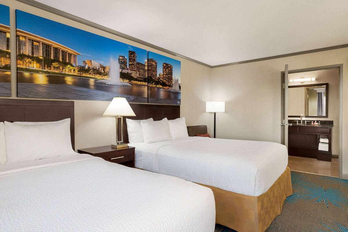 A hotel room with two beds, a nightstand with a lamp, a large window with a cityscape view, and a bathroom visible through an open door.