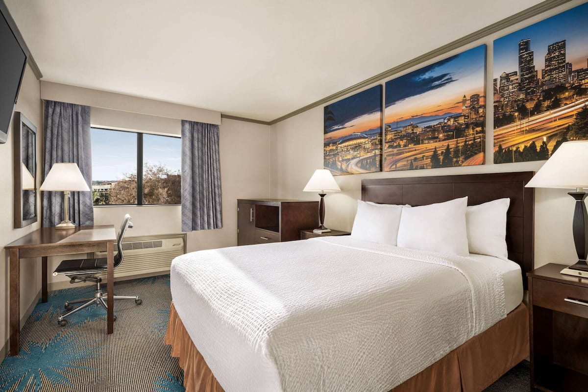 A hotel room with a large bed, a desk with a chair, a nightstand, and a window with curtains.