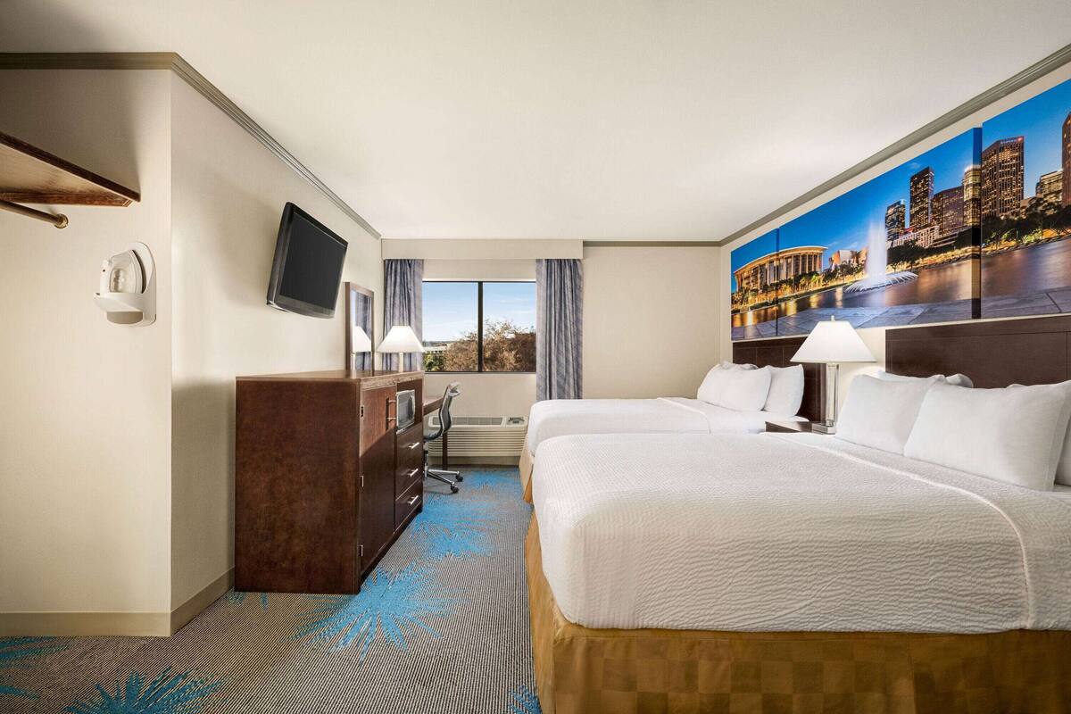 A hotel room with two beds, a desk with a chair, a television, and a large window with curtains.