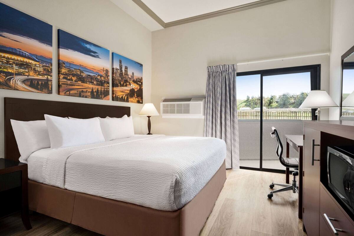 A hotel room with a large bed, a desk with a chair, a TV, and a balcony with a view of a cityscape.