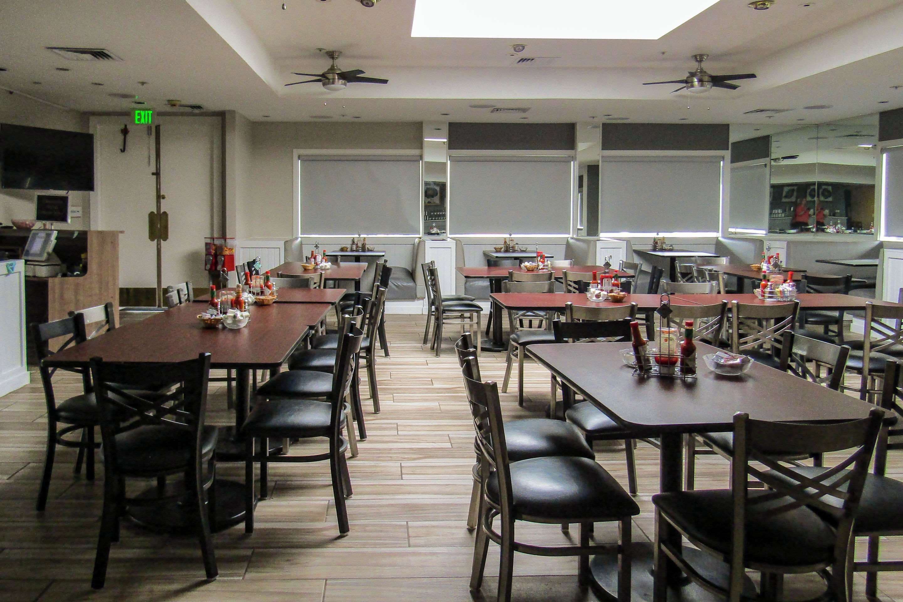 Un espace repas comprenant plusieurs tables et chaises, des ventilateurs de plafond et une vue sur la cuisine à travers les fenêtres.