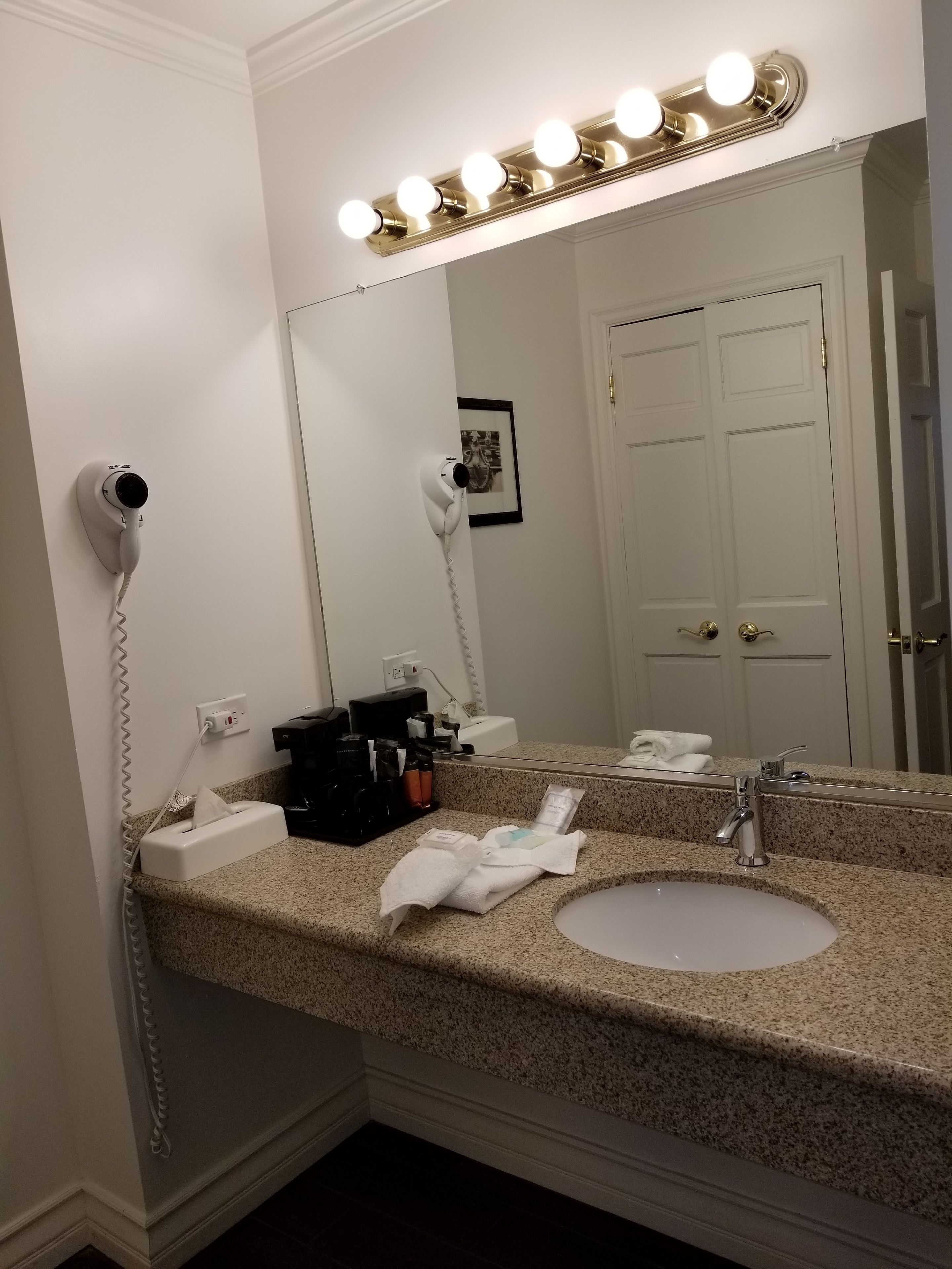 Une salle de bain d’hôtel avec un comptoir en granit, un lavabo, un miroir avec éclairage intégré, un sèche-cheveux et un porte-serviettes.
