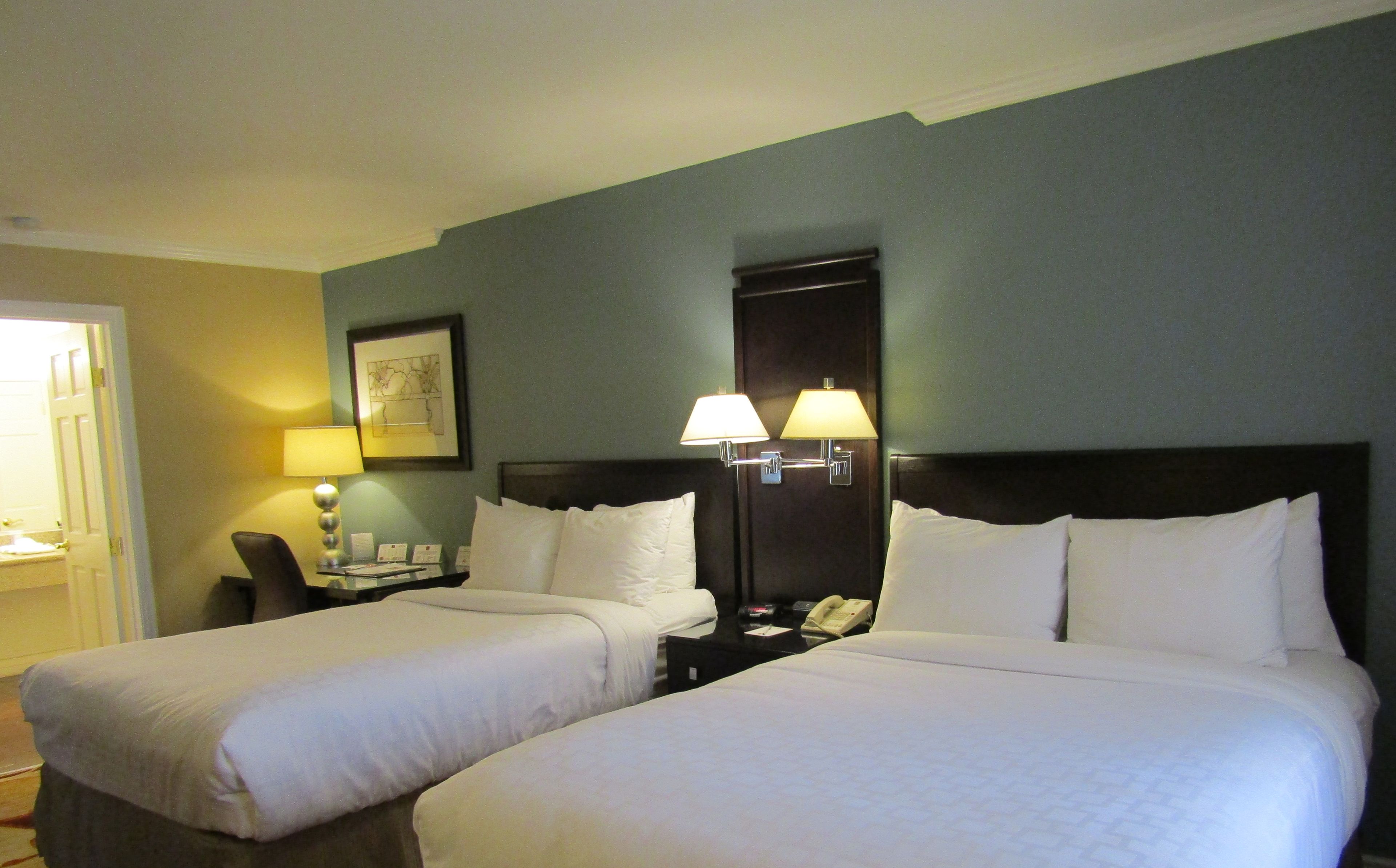 A hotel room with two beds, a nightstand with a lamp, a framed picture on the wall, and a door to the left.