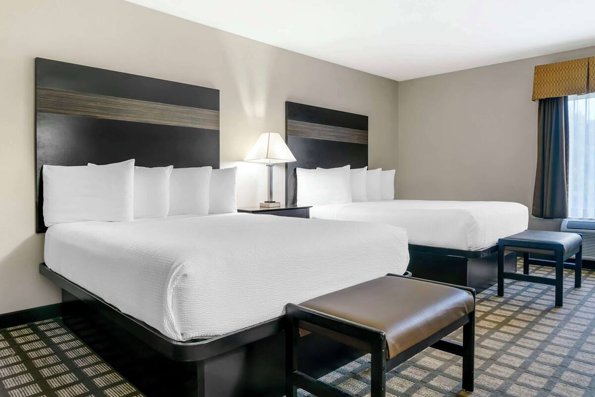 A hotel room with two beds, a nightstand, a lamp, and a bench.