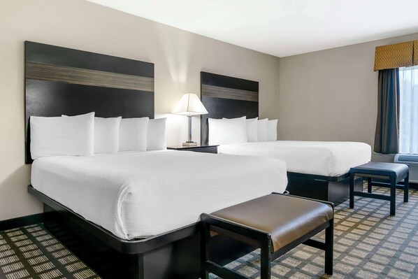 A hotel room with two beds, a nightstand, a lamp, and a bench.