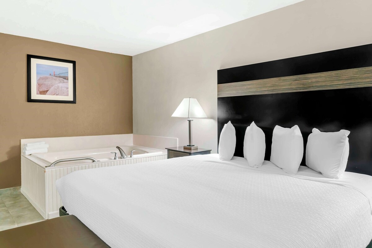 A hotel room with a large bed, a white bathtub, a nightstand with a lamp, and a framed picture on the wall.