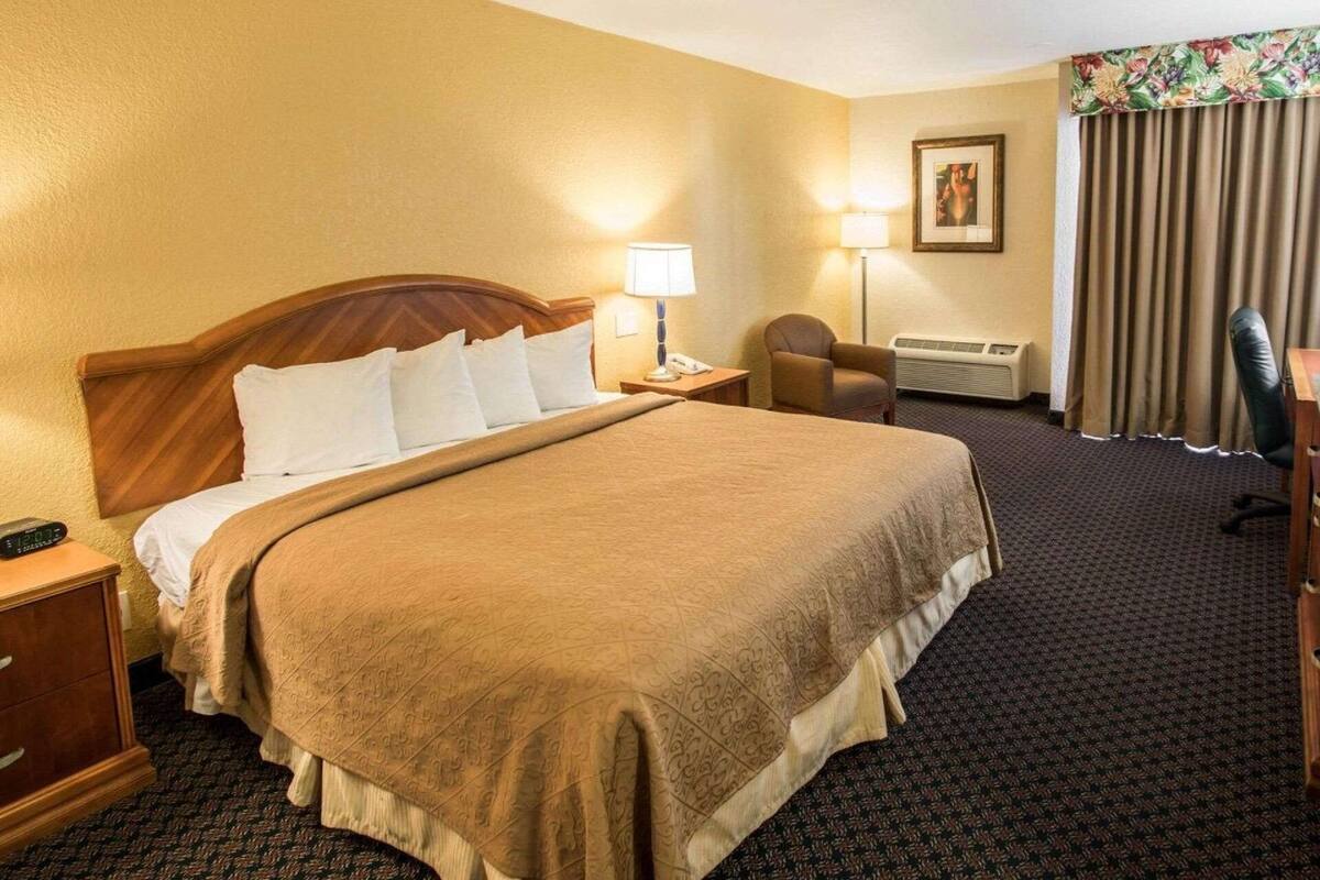 A hotel room with a large bed, a desk, a chair, a lamp, an air conditioner, and a framed picture on the wall.