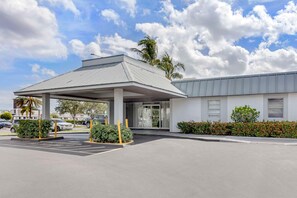 A hotel entrance with a covered parking area, a palm tree, and a building with a grey roof.