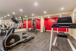 Academia com diversos equipamentos de exercício, incluindo esteiras e aparelhos de musculação, e uma parede vermelha com aviso de segurança.