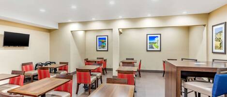 A hotel dining area with multiple tables and chairs, a flat-screen TV mounted on the wall, and framed pictures on the walls.