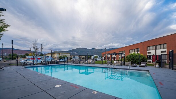 Una piscina con un borde de baldosas, un edificio de ladrillo al fondo y montañas a lo lejos.