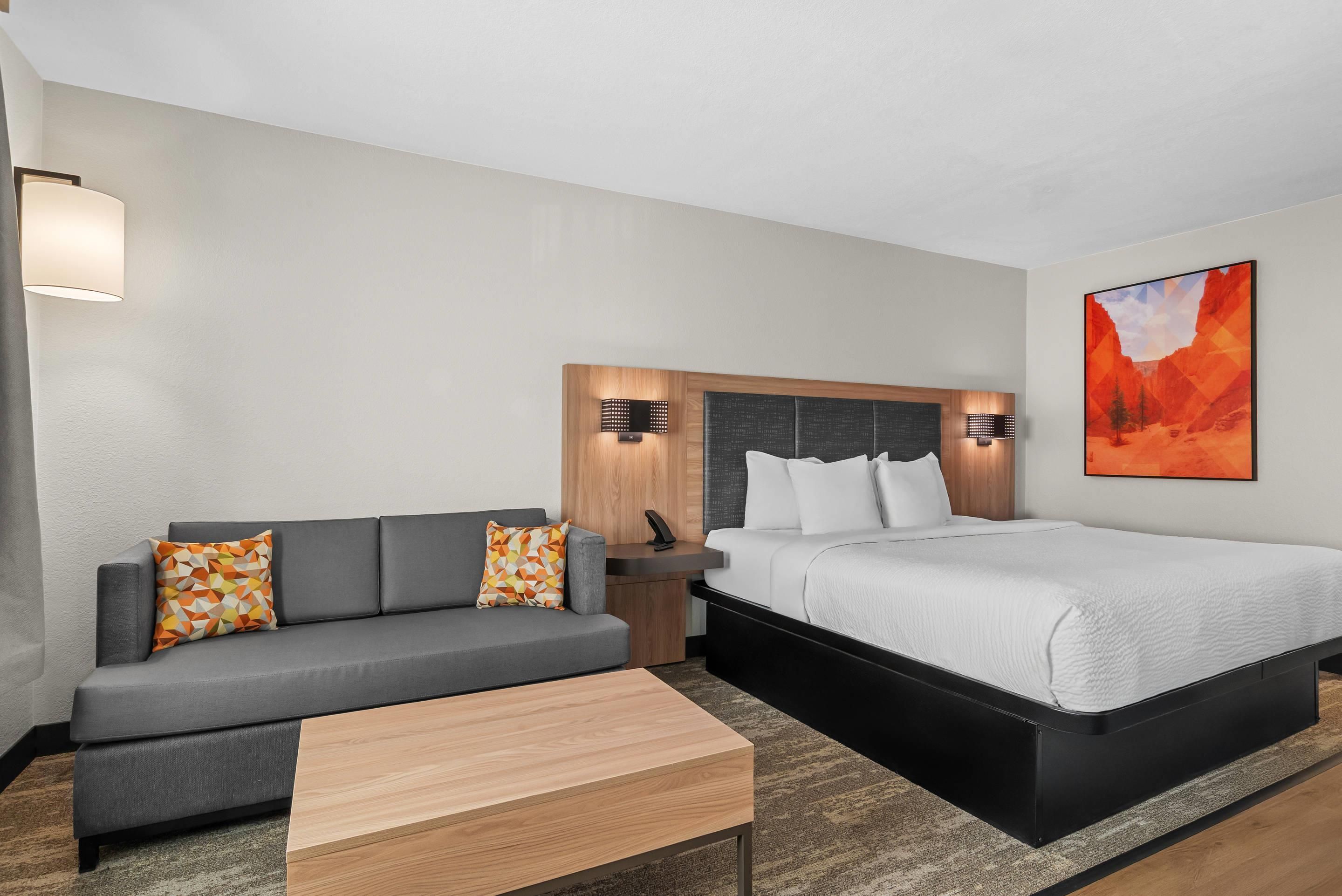 Una habitación de hotel con sofá, cama, mesita de noche, lámpara y un cuadro en la pared.