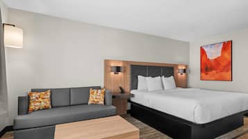 Una habitación de hotel con sofá, cama, mesita de noche, lámpara y un cuadro en la pared.