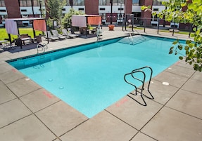 A rectangular pool with a clear blue water surface, surrounded by a tiled deck. There are lounge chairs and a small table with a chair, and a covered seating area with a table and chairs.