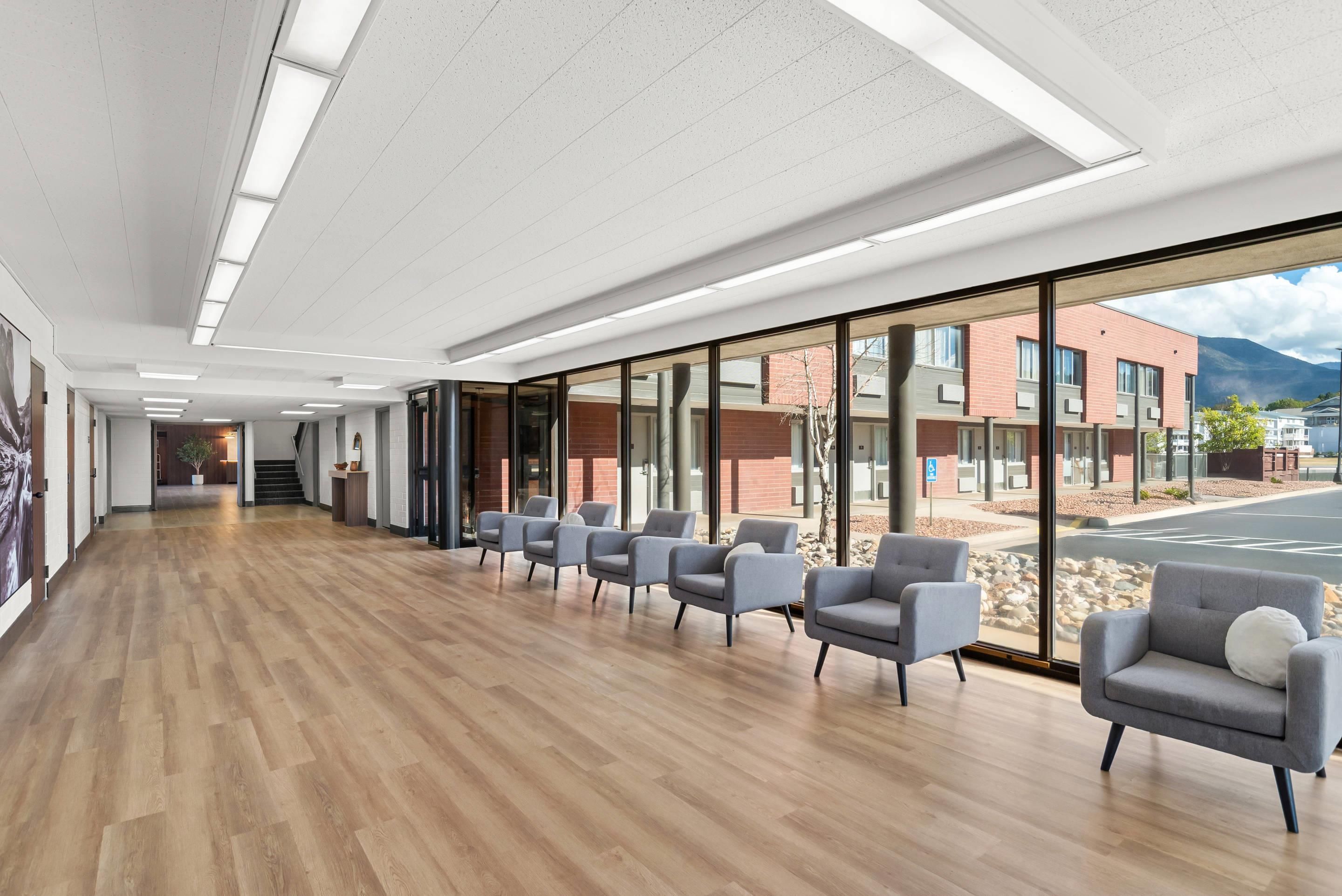 A modern waiting area with wooden flooring, grey chairs, and large windows offering a view of a building and mountains.
