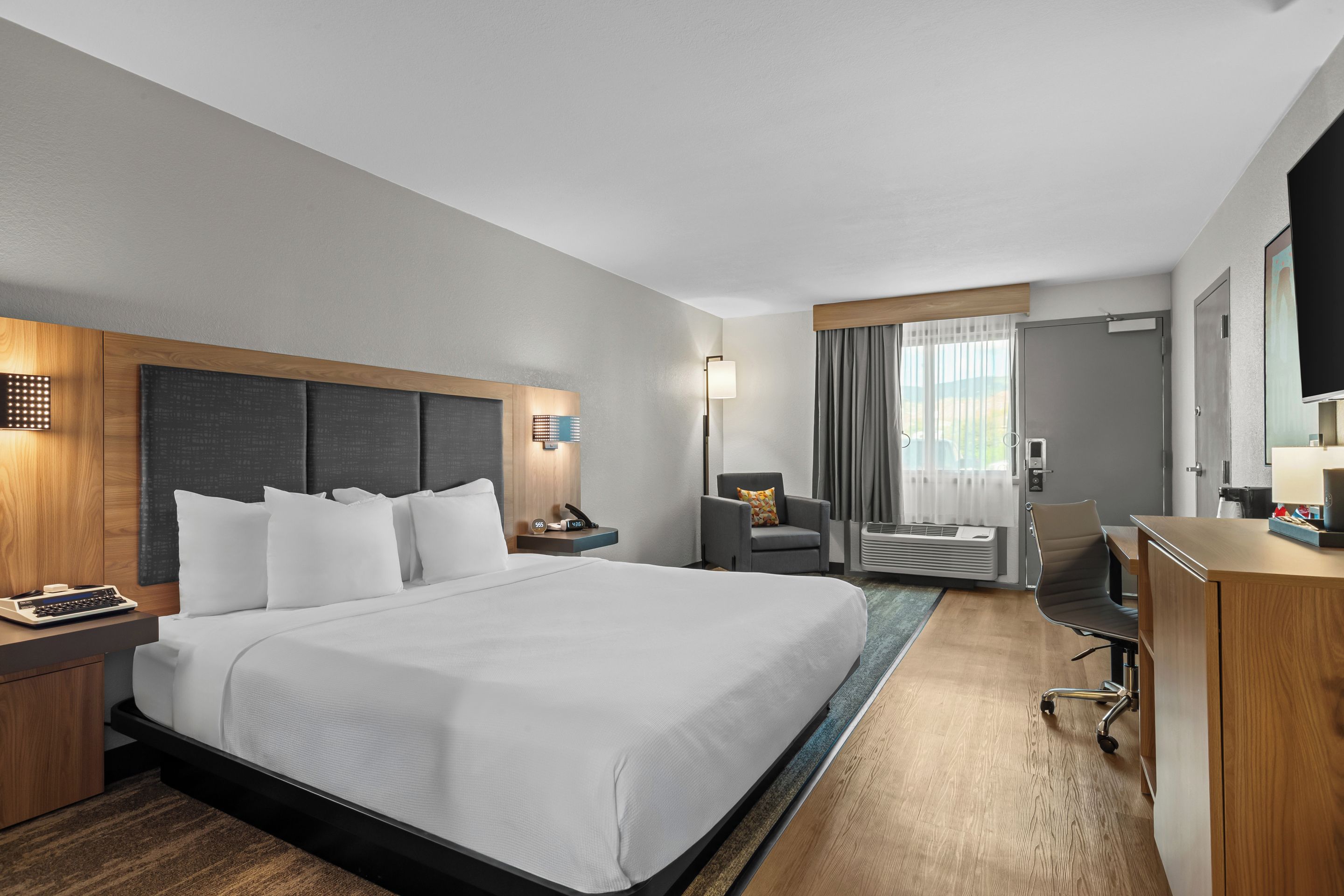 A hotel room with a large bed, a desk, a chair, a desk lamp, a nightstand, a TV, and a window with curtains.