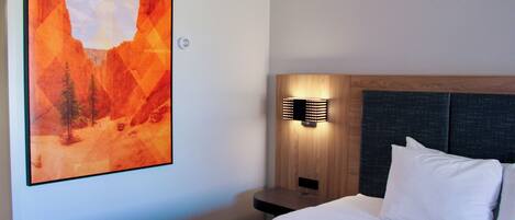 Una habitación de hotel con cama, mesita de noche, lámpara de pared y un cuadro grande de un paisaje montañoso.