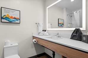 Une salle de bain moderne avec un lavabo blanc, un miroir et des toilettes. Un cadre contenant une citation est accroché au mur, ainsi qu’un porte-serviettes et un sac posé sur le comptoir.