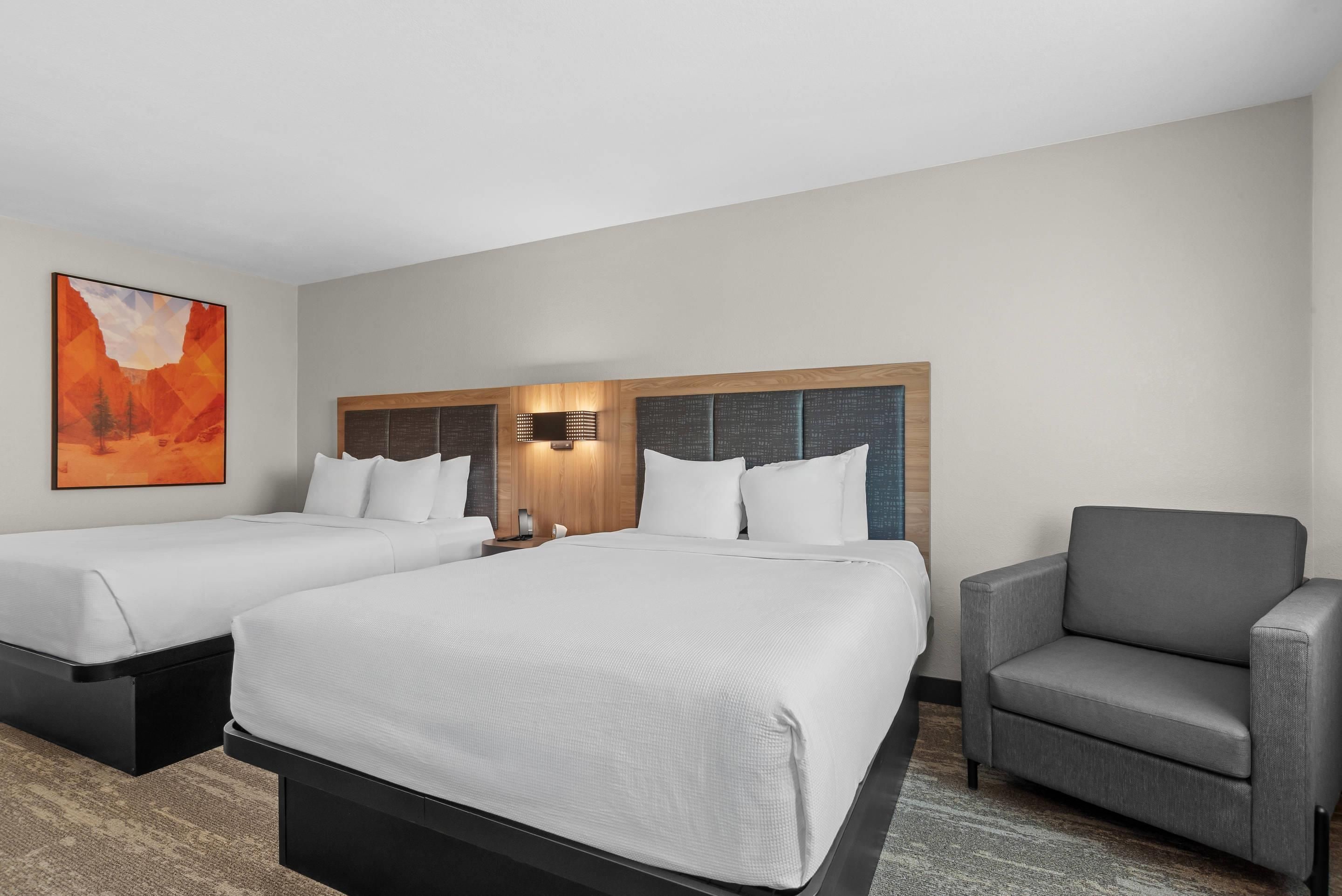 A hotel room with two beds, a gray armchair, and a large painting on the wall.