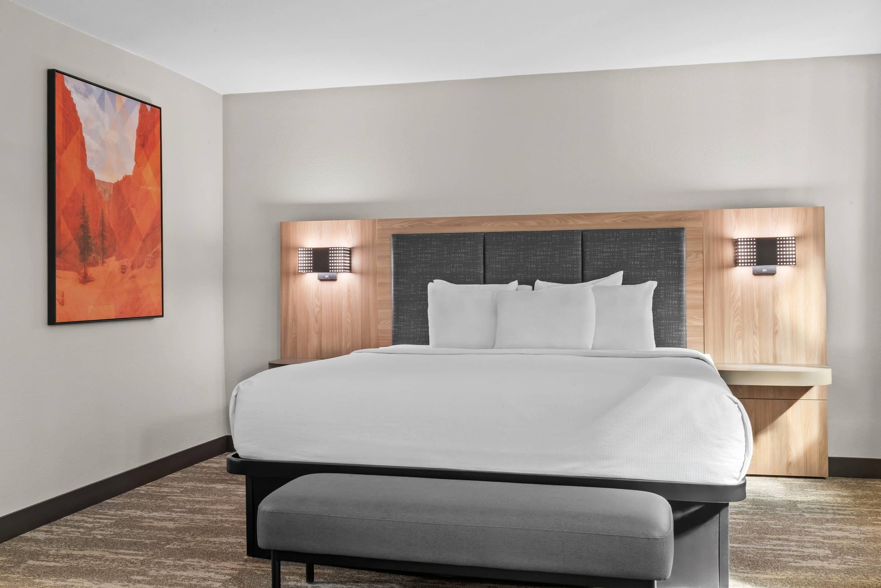 A modern hotel room with a large bed, a headboard with built-in lighting, a bench at the foot of the bed, and a painting on the wall.