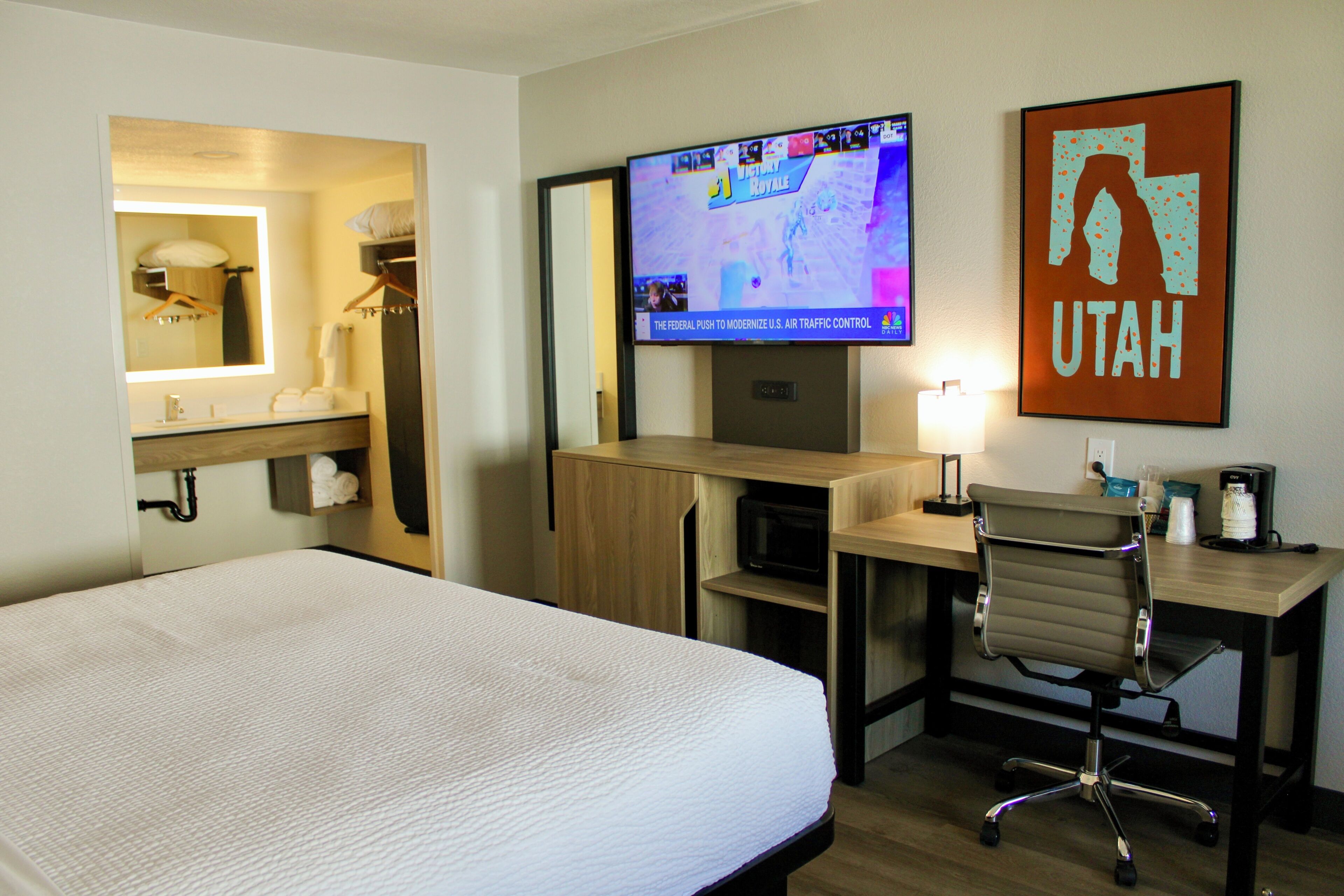 Ein Hotelzimmer mit einem Bett, einem Schreibtisch, einem Sessel, einem Fernseher und einem gerahmten Schild an der Wand.