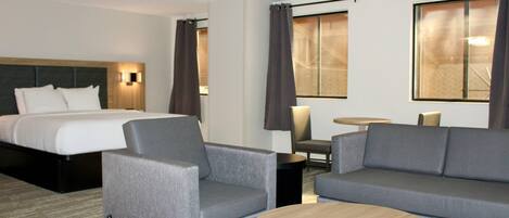 Ein Hotelzimmer mit einem Bett, zwei grauen Sofas, einem runden Holztisch und einem kleinen Tisch mit einer Lampe.