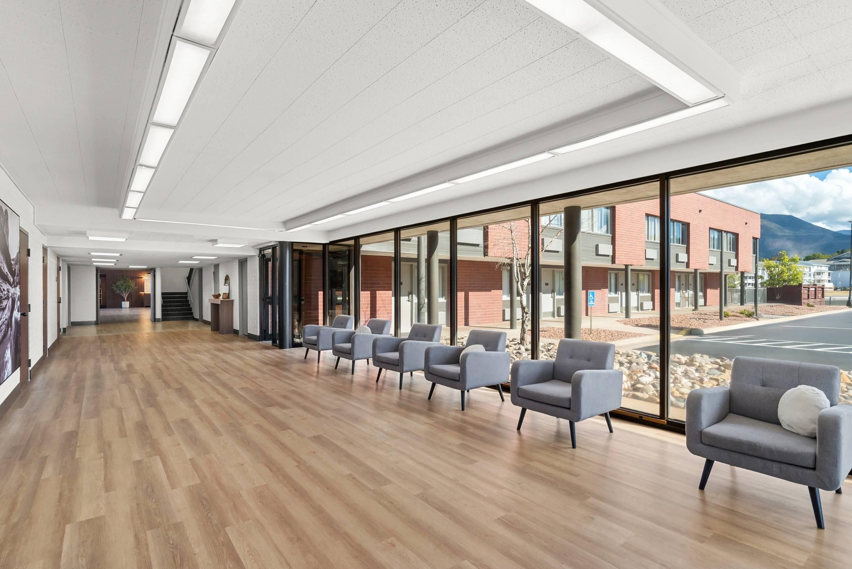 A modern waiting area with a wooden floor, grey chairs, and large windows offering a view of a building and mountains.