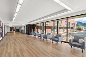 A modern waiting area with a wooden floor, grey chairs, and large windows offering a view of a building and mountains.
