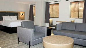 A hotel room with a bed, two grey armchairs, a grey sofa, a round wooden coffee table, a small round table with a lamp, and two windows with curtains. - The Cairn Hotel, an Ascend Collection Hotel (Cedar City)