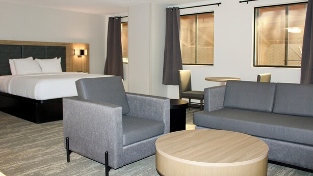 Habitación de hotel con una cama, dos sillones grises, un sofá gris, una mesa de centro redonda de madera, una pequeña mesa redonda con una lámpara y dos ventanas con cortinas.