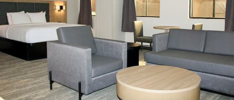 A hotel room with a bed, two grey armchairs, a grey sofa, a round wooden coffee table, a small round table with a lamp, and two windows with curtains.