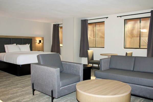 Ein Hotelzimmer mit einem Bett, zwei grauen Sesseln, einem grauen Sofa, einem runden Holztisch, einem kleinen runden Tisch mit Lampe und zwei Fenstern mit Vorhängen.