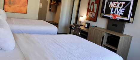 A hotel room with two beds, a TV displaying 'NEXT LIVE,' a mirror, and a desk with a lamp.