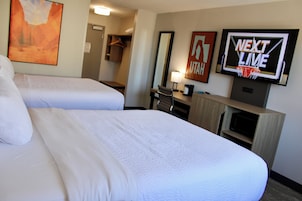Habitación de hotel con dos camas, un televisor mostrando 'NEXT LIVE', un espejo y un escritorio con una lámpara.