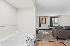 Een moderne hotelkamer met een ruime badkuip, een zithoek met een bank en een tafeltje, en uitzicht op het omliggende landschap door de ramen.