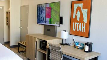 A hotel room with a desk, chair, television, coffee maker, and a framed sign.