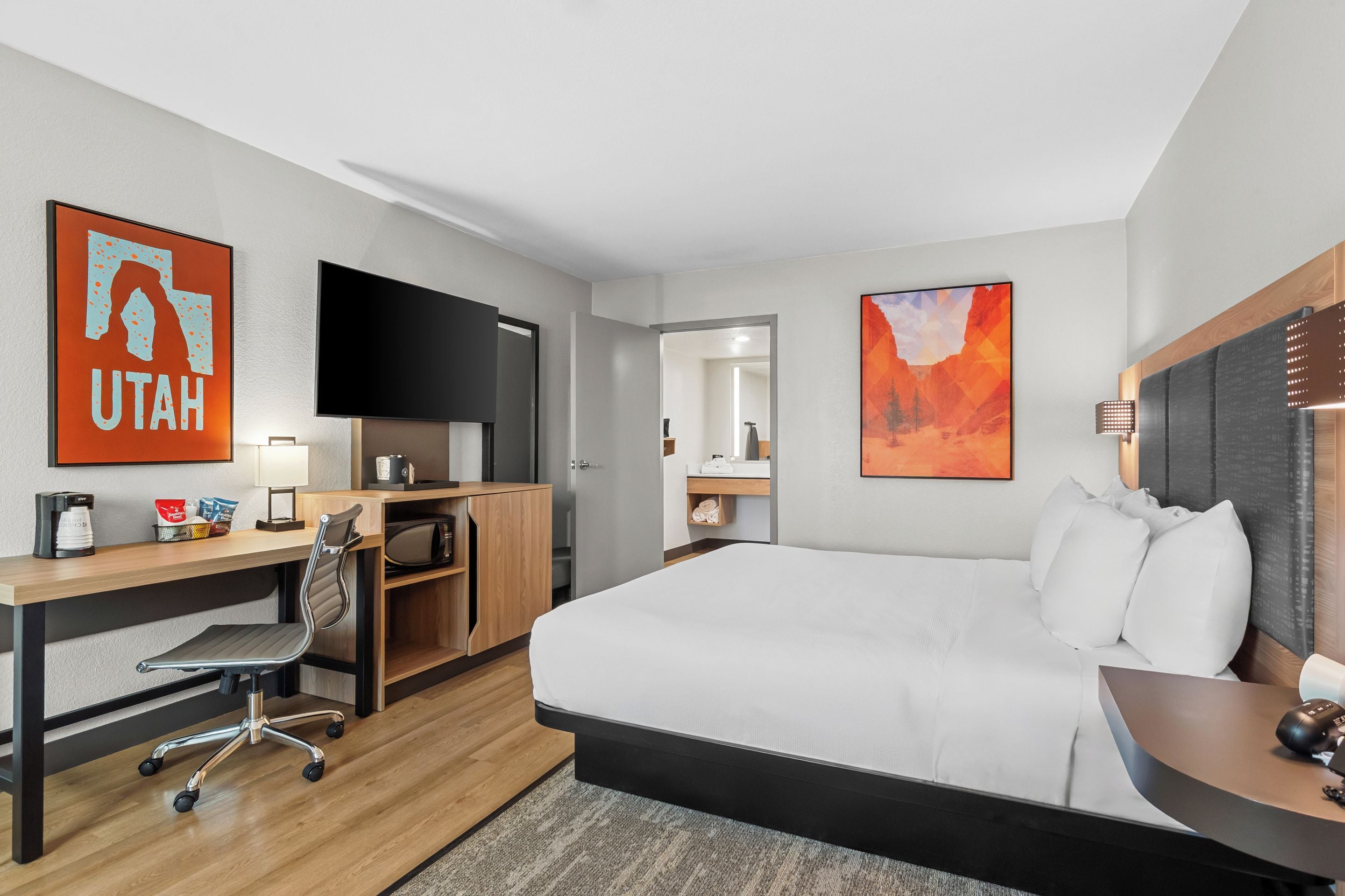 A hotel room with a bed, desk, chair, TV, artwork, and a bathroom visible through an open door.