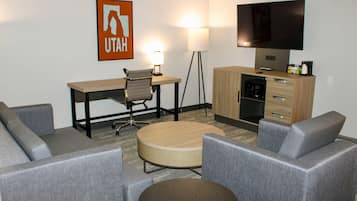 A hotel room with a sofa, a round coffee table, a desk, a lamp, a television, and a kitchen area.