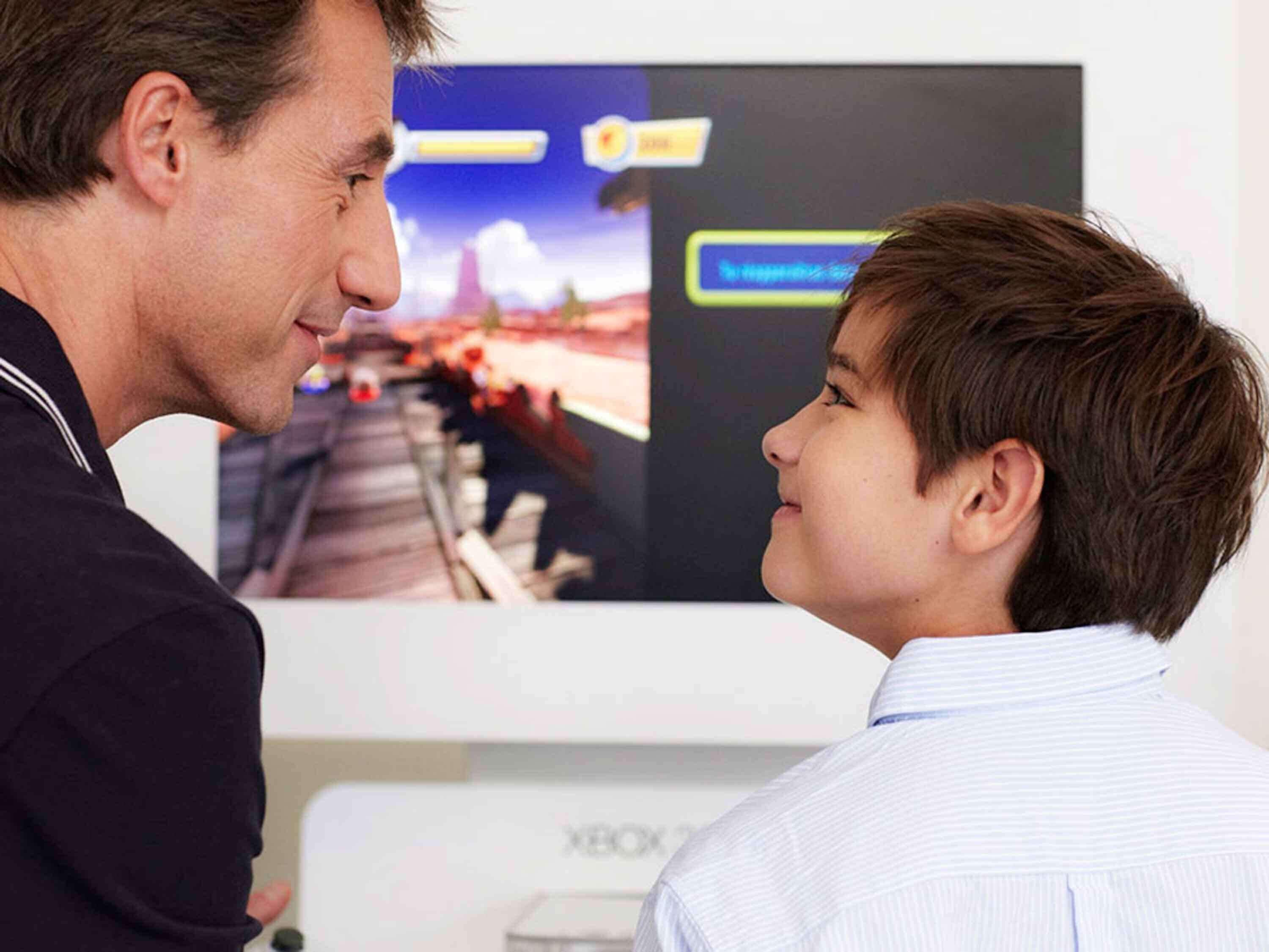 A person in a dark jacket and another in a light striped shirt facing a large screen displaying a video game scene.