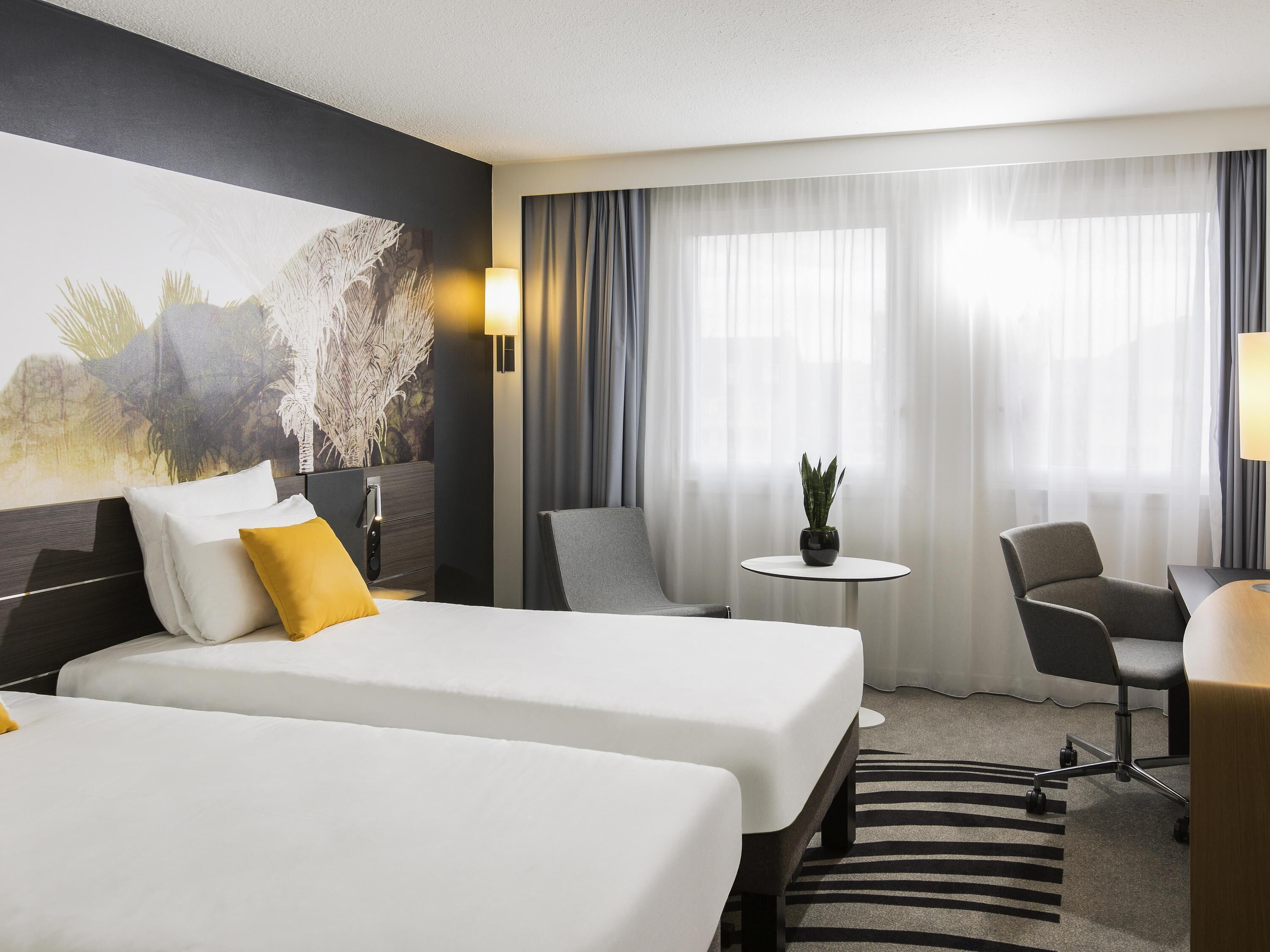 Une chambre d’hôtel avec deux lits, un bureau, une chaise, une petite table et une grande fenêtre avec des rideaux.