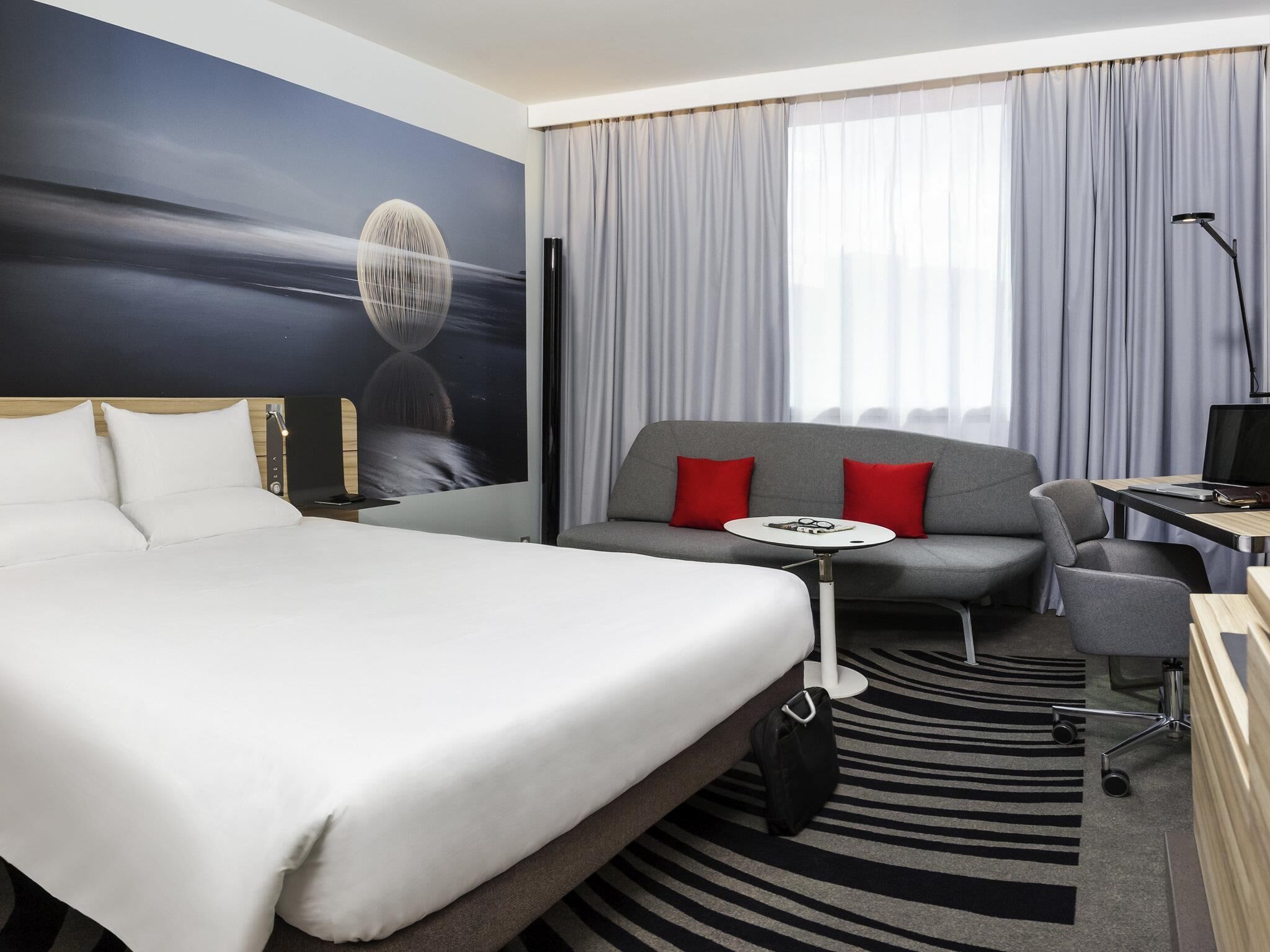 A hotel room with a bed, a sofa, a small table, a desk, a chair, a lamp, and a large window with curtains.