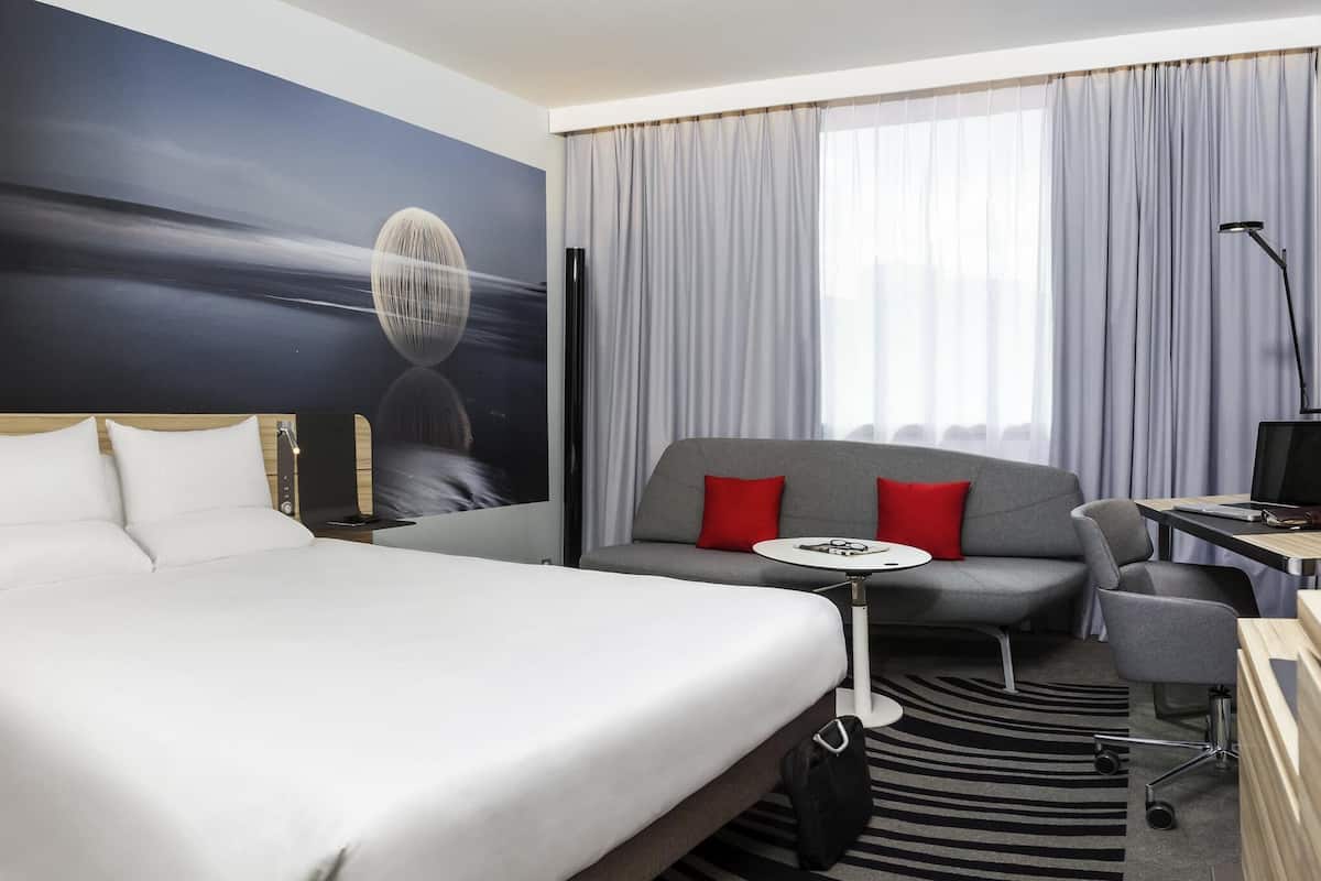 A hotel room with a bed, a sofa, a small table, a desk, a chair, a lamp, and a large window with curtains.