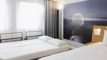 A hotel room with two beds, a large window with curtains, a wall-mounted artwork, and a bedside table with a lamp.