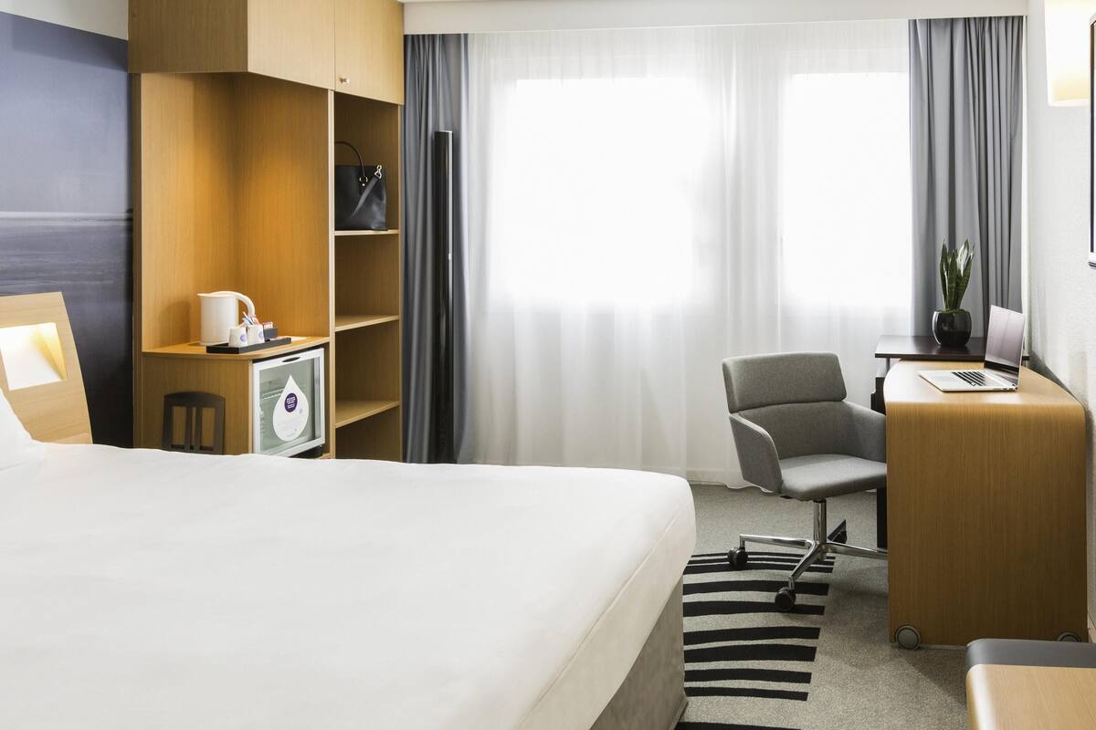 A hotel room with a bed, a desk with a laptop, a chair, a small TV, and a bookshelf.