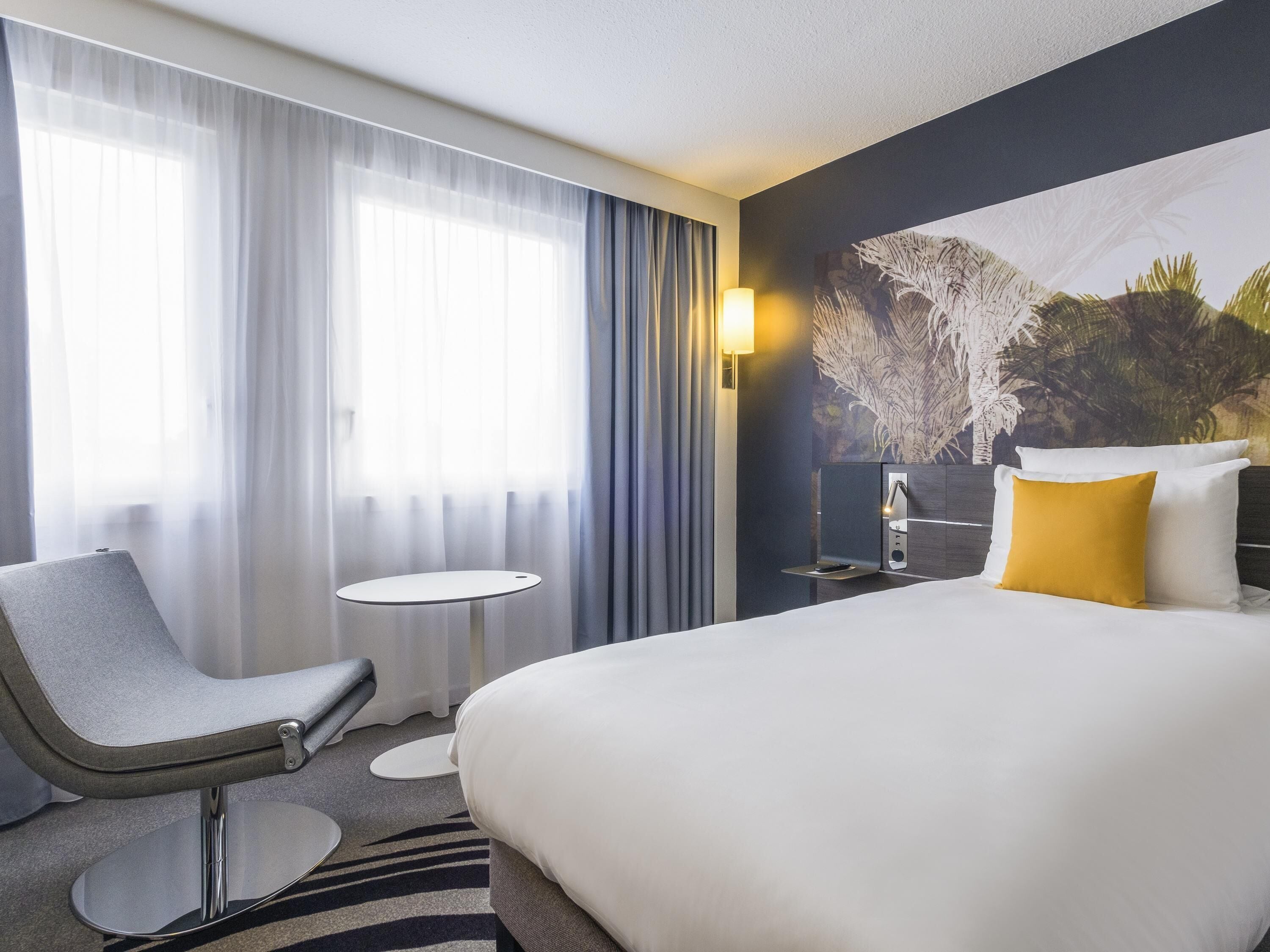 A hotel room with a bed, a chair, a small table, a nightstand, and a wall-mounted lamp.