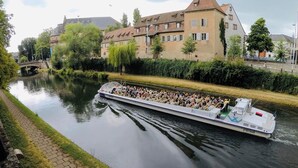 A riverboat with passengers, surrounded by buildings and greenery. - Novotel Strasbourg Centre Halles (Strasbourg)