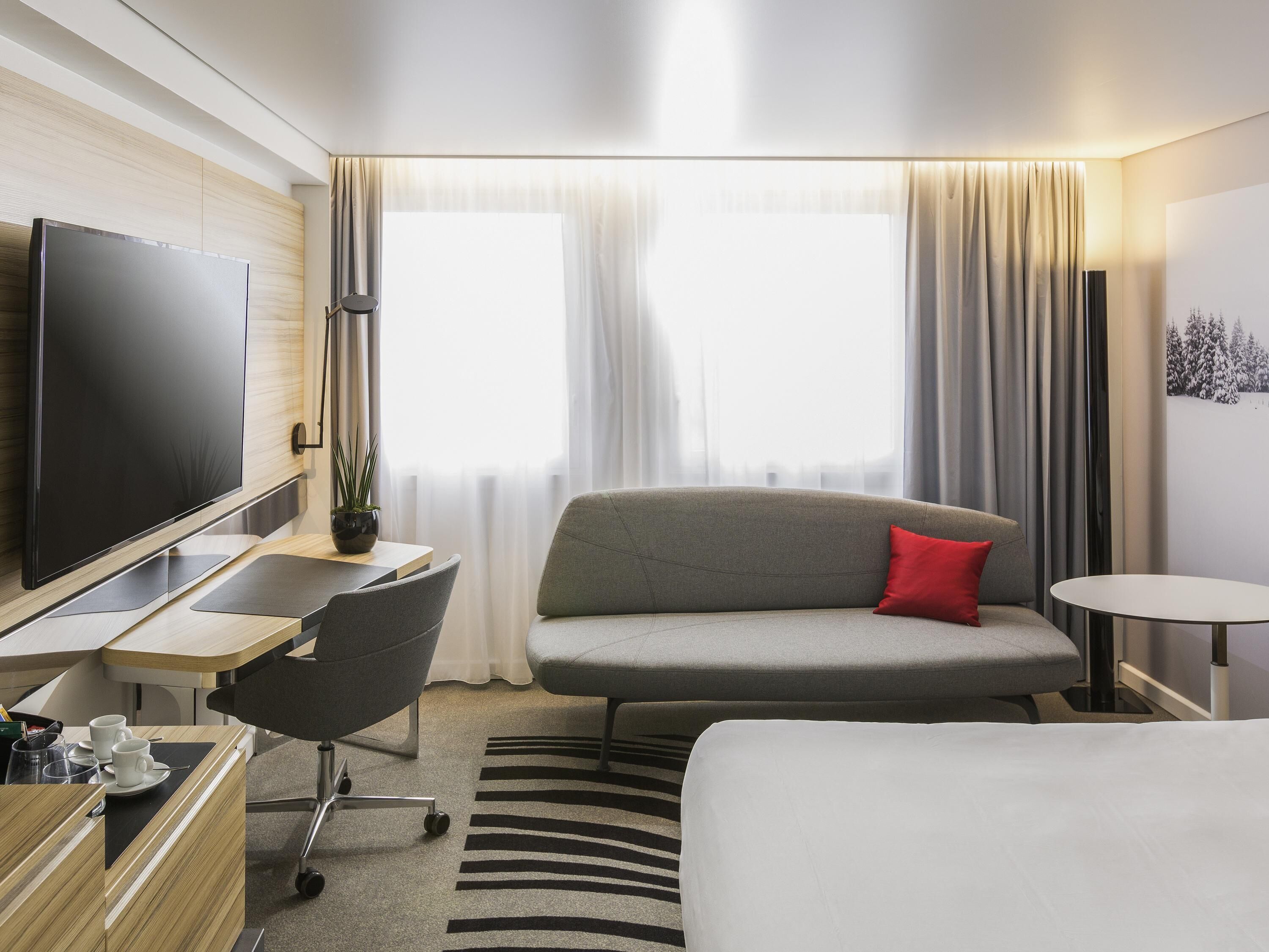 A modern hotel room with a flat-screen TV, a sofa with a red pillow, a small round table, a desk with a chair, and a bed.