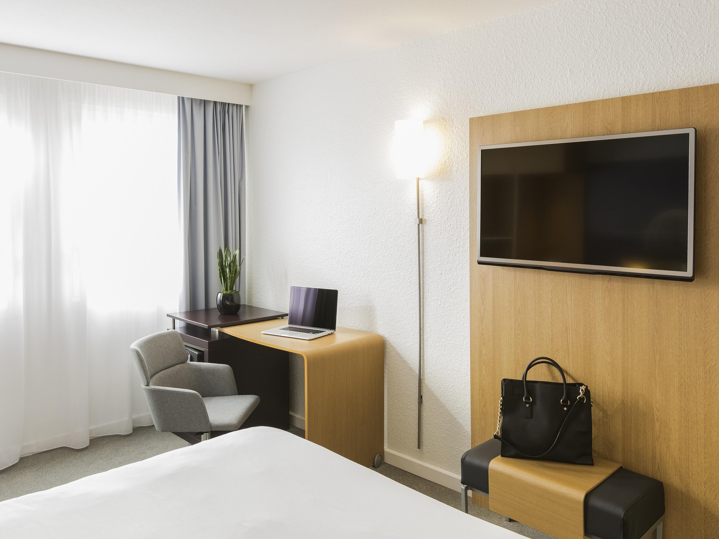 Quarto de hotel com cama, escrivaninha com um laptop, cadeira, televisão na parede, candeeiro de chão e uma bolsa em um banco.