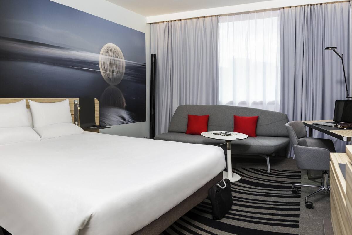A hotel room with a bed, a sofa, a desk, a chair, a small table, and a large window with curtains.